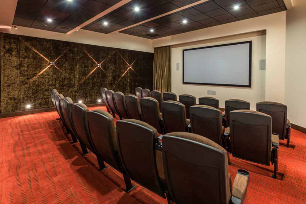 A small private theater room with rows of black seats facing a large blank screen, red carpet flooring, dark textured walls, and modern wall lights creating a cozy atmosphere.