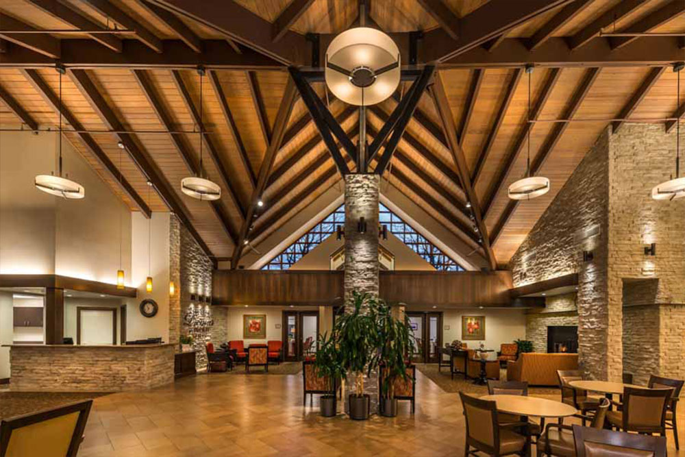 Spacious hotel lobby with high wooden vaulted ceiling, exposed beams, stone pillars, modern chandeliers, comfortable seating, large plant centerpiece, and warm ambient lighting. Reception desk is visible to the left.