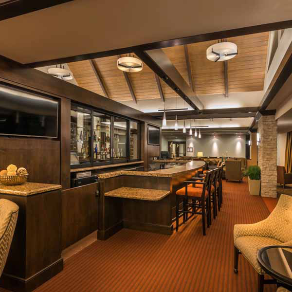 A modern hotel bar and lounge with wooden beams, pendant lights, a large bar counter with high chairs, a TV, and a seating area with upholstered chairs on a red carpet. Bottles are displayed behind the bar.