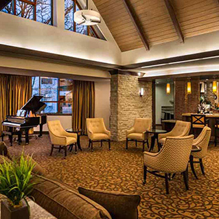 Spacious, warmly lit lounge with a grand piano, cushioned chairs around small tables, patterned carpet, large windows with curtains, stone accent walls, and a vaulted wooden ceiling.