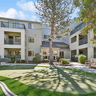 A small putting green with holes sits in the courtyard of a modern apartment complex, surrounded by greenery, balconies, and large windows under a sunny sky.