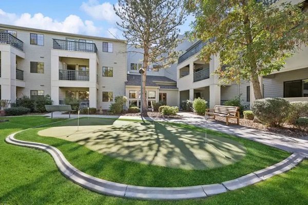 A courtyard features a small putting green surrounded by manicured grass, shrubs, a tree, a wooden bench, and a multi-story apartment building with balconies in the background under a partly cloudy sky.
