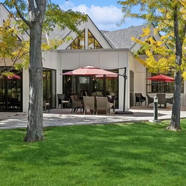 A modern white building with large windows and a patio featuring tables, chairs, and red umbrellas. Green grass and trees with yellow leaves are in the foreground under a blue sky.