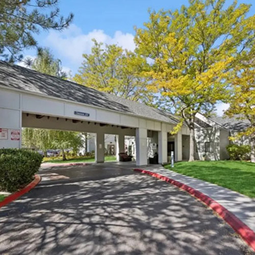 A building with a covered entrance driveway, surrounded by green grass, bushes, and trees with yellow-green leaves under a blue sky. A red curb lines the driveway.