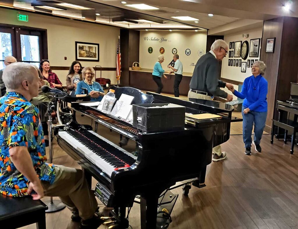 A group of seniors in a community room enjoying a live piano performance. Some are sitting and smiling, while others dance together. The pianist wears a colorful shirt, and the atmosphere is lively and cheerful.