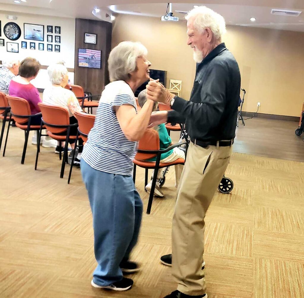 An elderly man and woman smile joyfully as they dance together in a community room, while other seniors sit and watch in the background. The room has warm lighting and framed photos on the wall.