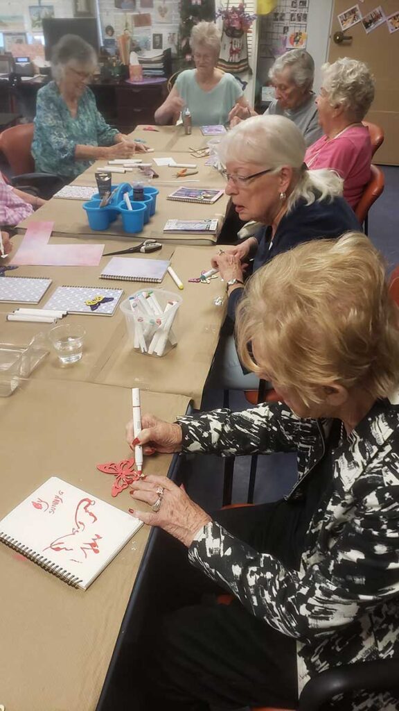A group of elderly women sit around a table in a classroom, engaging in arts and crafts activities with markers, paper, and supplies. They appear focused and are working on creative projects together.