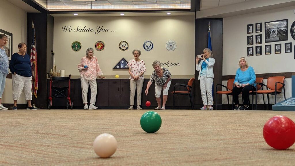 A group of older women play an indoor game of bocce ball in a community room. Some are standing while others sit or watch. Military emblems and a “We Salute You” sign are displayed on the back wall.