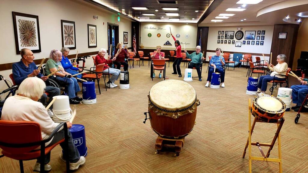 A group of seniors sits in a circle in a community room, each playing drums or buckets. The atmosphere is lively and engaging, with chairs arranged in a circle and drums in the center of the room.