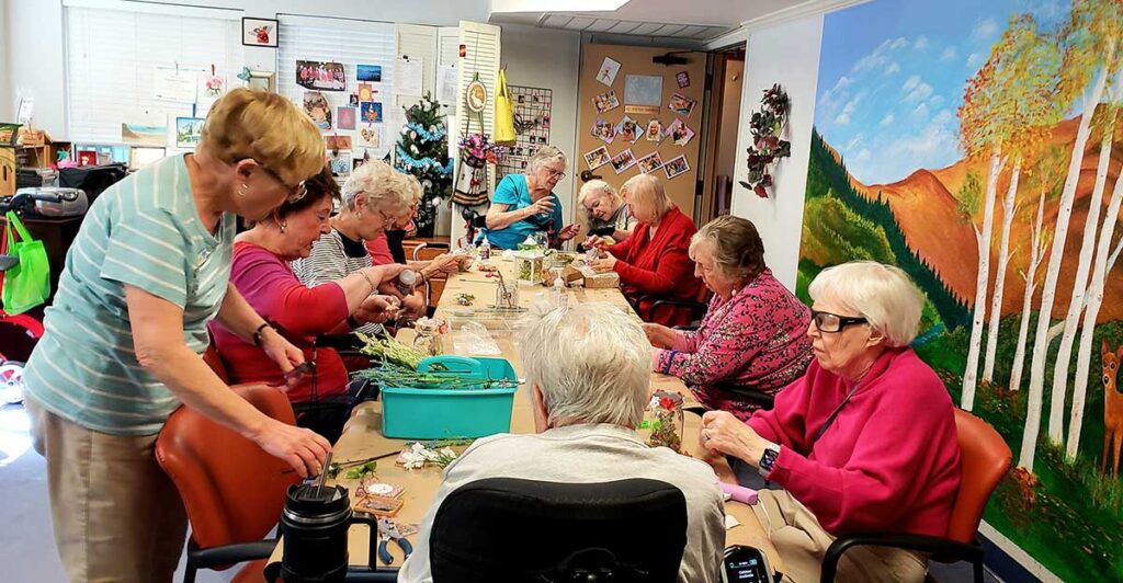 A group of elderly women sit around a long table in a bright room, engaging in arts and crafts activities. Supplies are scattered on the table, and colorful artwork decorates the walls.