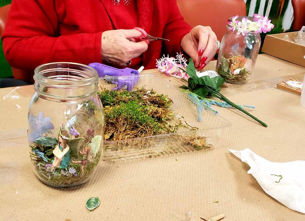 A person in a red shirt is crafting a fairy garden inside a glass jar using moss, artificial flowers, and small figurines. Craft materials and tools are spread out on a table.