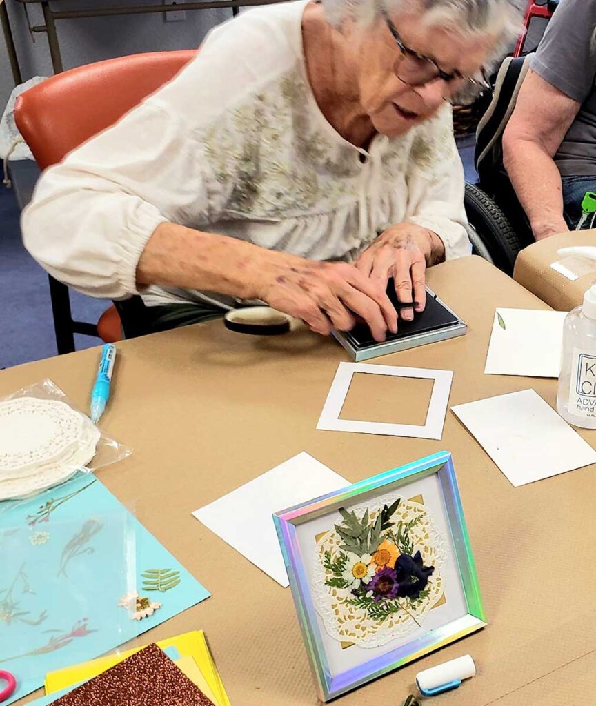 An elderly woman wearing glasses works on a craft project at a table, surrounded by paper, art supplies, and a colorful framed piece featuring pressed flowers.