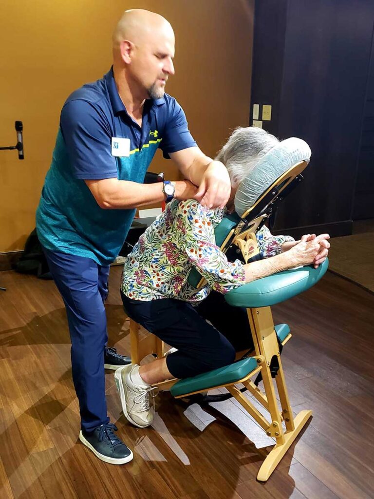 A man gives a seated massage to an older woman who is leaning forward in a padded massage chair. He is pressing on her shoulder while she rests her arms on the chair, in a room with wooden flooring.