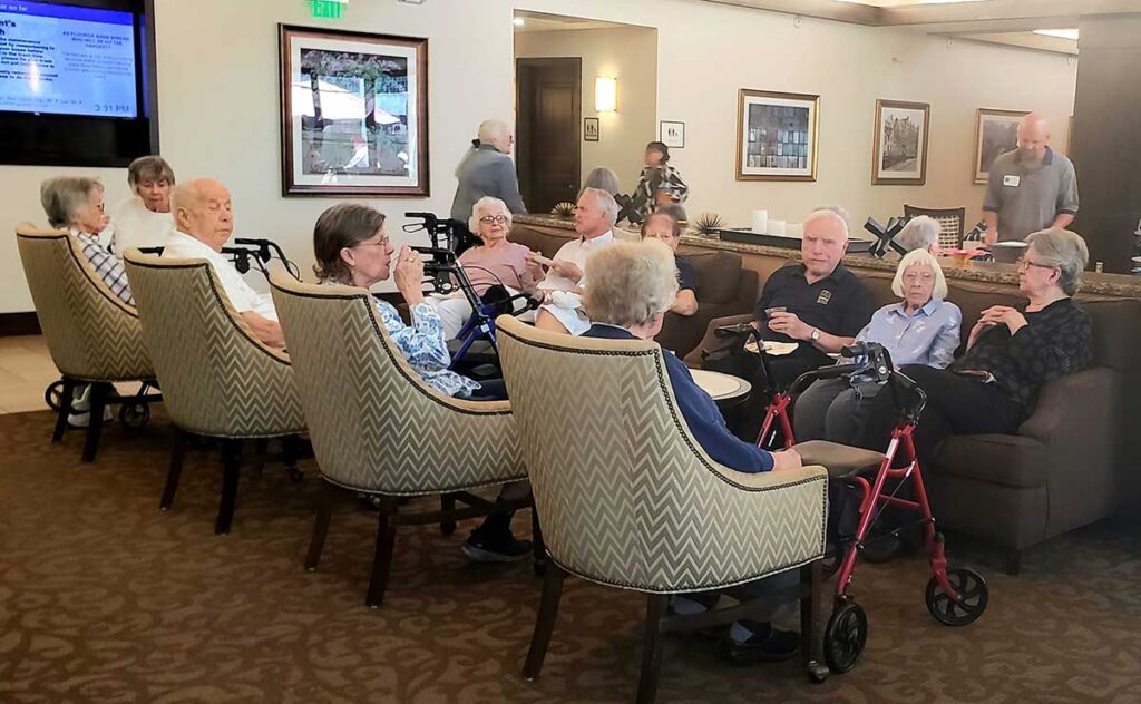 A group of elderly people sits in armchairs and on sofas in a lounge area, conversing and relaxing. Some use walkers. The room is warmly lit with framed art on the walls and other people visible in the background.