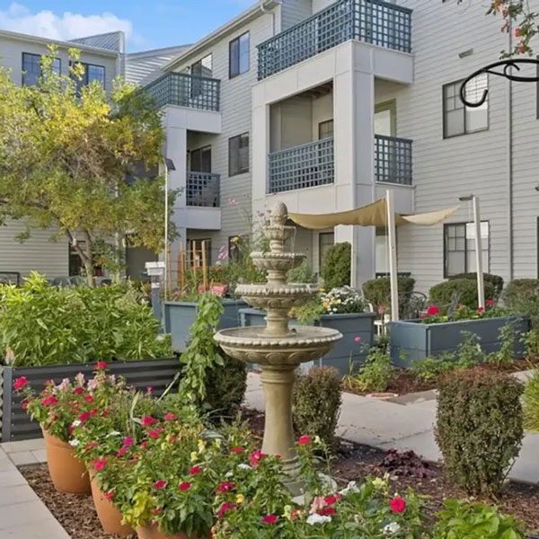 A courtyard of an apartment complex featuring a tiered stone fountain, potted flowers, raised garden beds, green shrubs, and balconies on the white building in the background.
