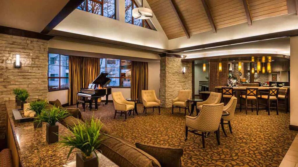 A spacious, elegant lounge with upholstered chairs, a grand piano by large windows, pendant lights above a bar, decorative plants, stone accents, and a high, vaulted ceiling with exposed beams.