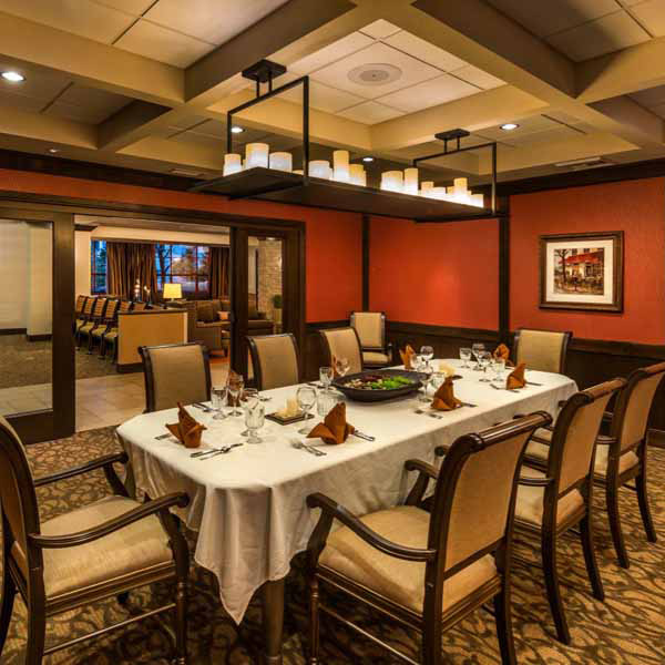 A formal dining room with a rectangular table set for eight, elegant chairs, folded napkins, wine glasses, and warm lighting. The room has red walls, framed artwork, and a view into an adjacent lounge area.