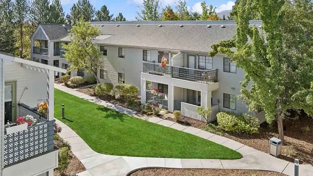 Two-story apartment buildings with balconies overlook a landscaped courtyard with green grass, trees, bushes, and winding concrete walkways on a bright, sunny day.