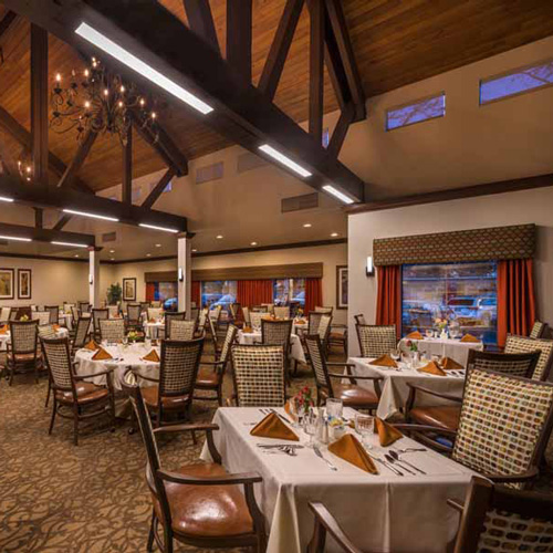 A spacious, elegant restaurant dining room with wooden beams, chandeliers, neatly set tables with white cloths, and brown napkins. Large windows and warm lighting create a cozy atmosphere.