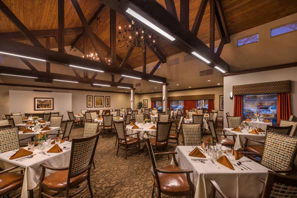 A spacious, elegant restaurant dining room with many tables set with white tablecloths, brown napkins, and place settings. Wooden chairs surround the tables, and the ceiling features exposed beams and chandeliers.