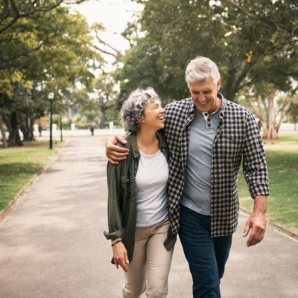An older couple walks arm in arm down a tree-lined park path, smiling and enjoying each other's company on a pleasant day.