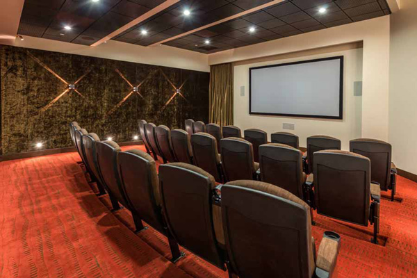 A small home theater room with rows of black cushioned seats, a red carpet, a large blank projection screen on the front wall, and dim wall lights creating a cozy ambiance.