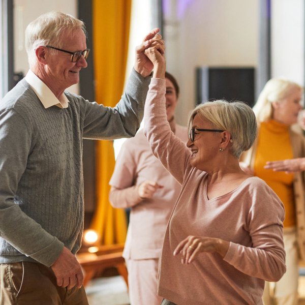An older man and woman smile and dance together indoors, holding hands. Other people are visible in the background, and the atmosphere looks joyful and relaxed.