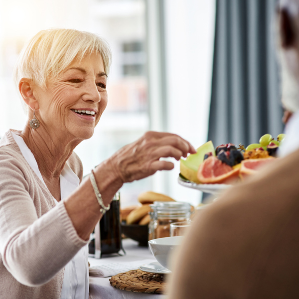 Smiling older woman with short gray hair reaches for a plate of fruit, including grapefruit and grapes, while sitting at a dining table in a bright, cozy room.