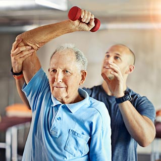 An older man in a blue polo shirt lifts a small red dumbbell with one arm, assisted by a younger man in a gym setting. The younger man is providing support and guidance.