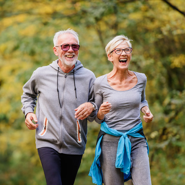 An older man and woman, both wearing glasses and casual athletic clothes, are smiling and jogging outdoors together on a tree-lined path.