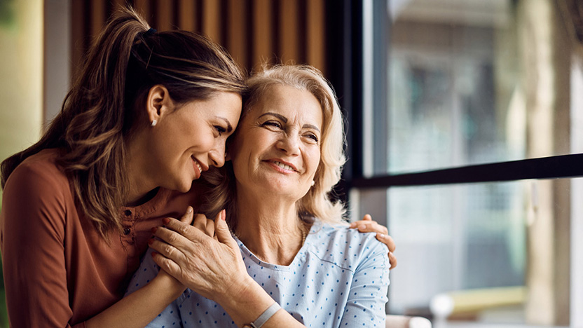 Two women sit closely together, smiling and embracing near a window. Their warmth and affection highlight the importance of family involvement in senior care within a well-lit, cozy setting.