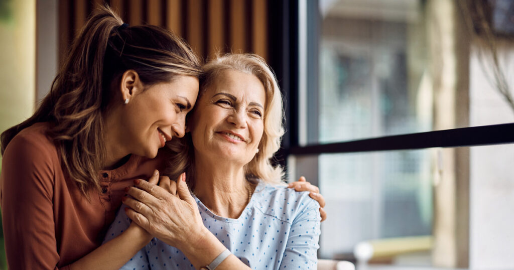 A young woman lovingly hugs an older woman from behind, both smiling and sitting near a window with natural light streaming in. The scene conveys warmth, affection, and a strong emotional bond.