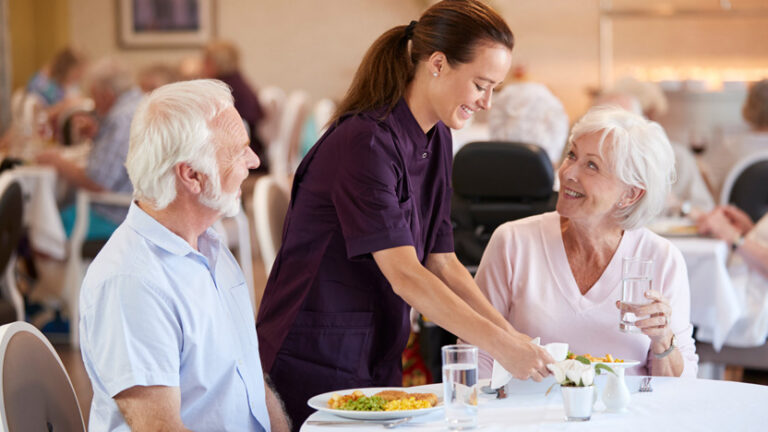 A young female server brings food to a smiling elderly couple seated at a table in a bright, busy dining room, where family involvement in senior care helps create a warm and engaging atmosphere.