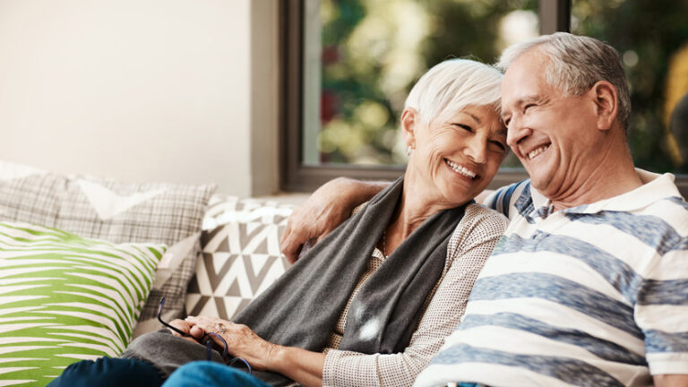 An elderly couple sits closely on a sofa, smiling and laughing together. The man has his arm around the woman as they enjoy the bright natural light coming through a window behind them. Pillows with geometric patterns are on the couch, creating a cozy atmosphere that reflects the comfort and financial advantages of independent living.